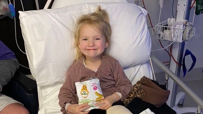 A smiling blonde toddler clutches a bag of sweets while she lies in a hospital bed.