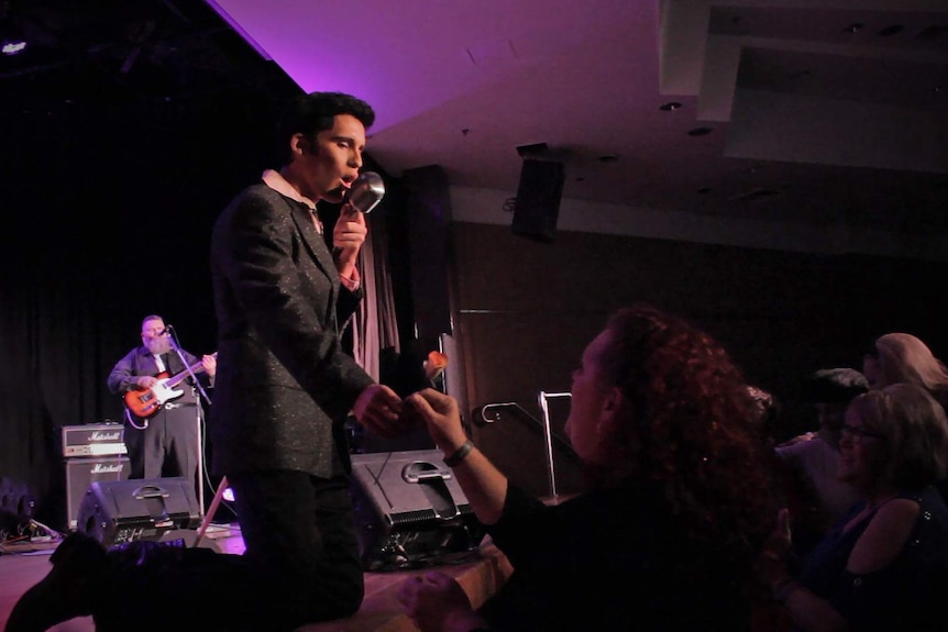 Elvis tribute artist Brody Finlay kneels on stage while giving a lady in the audience a rose