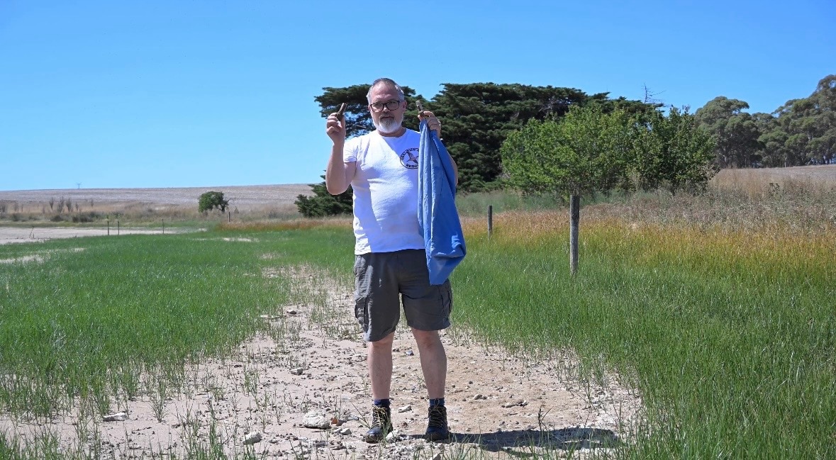 Un hombre con barba y gafas sostiene dos cartuchos de escopeta y una bolsa de basura.