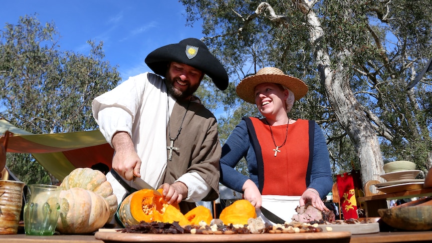 A man in medieval dress cuts pumpkin beside a woman slicing meat, under a blue sky.