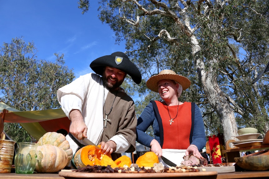 A man in medieval dress cuts pumpkin beside a woman slicing meat, under a blue sky.