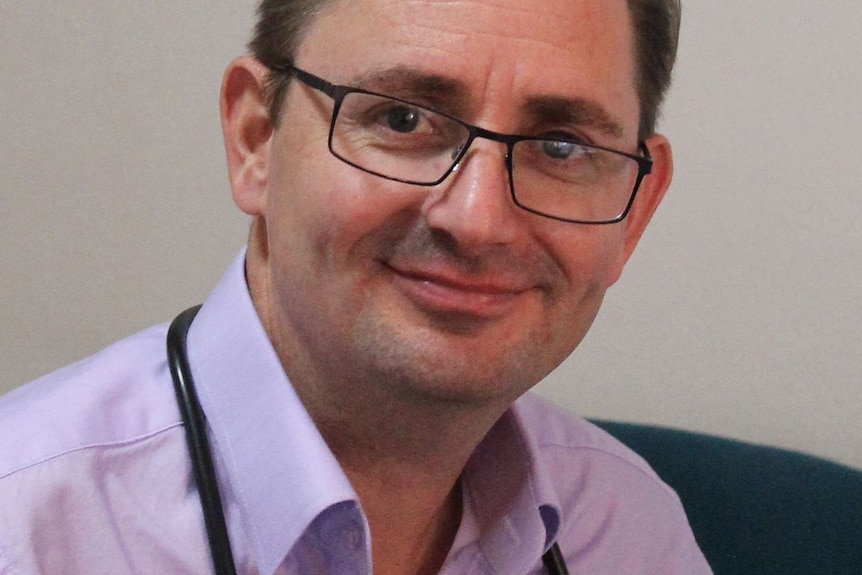 A man in a purple shirt and glasses with a stethoscope around his neck smiles as he sits at a desk.