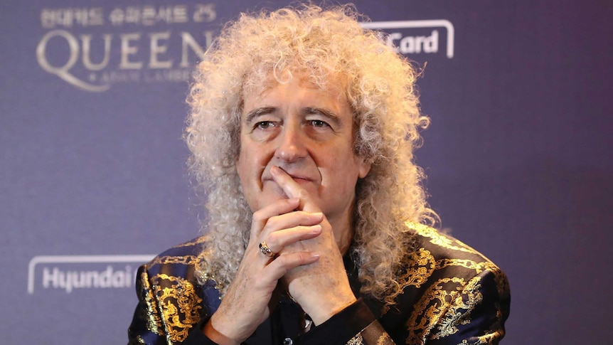 A slim man with frizzy white hair in a sequined jacket puts his hands to his face seated at a table.