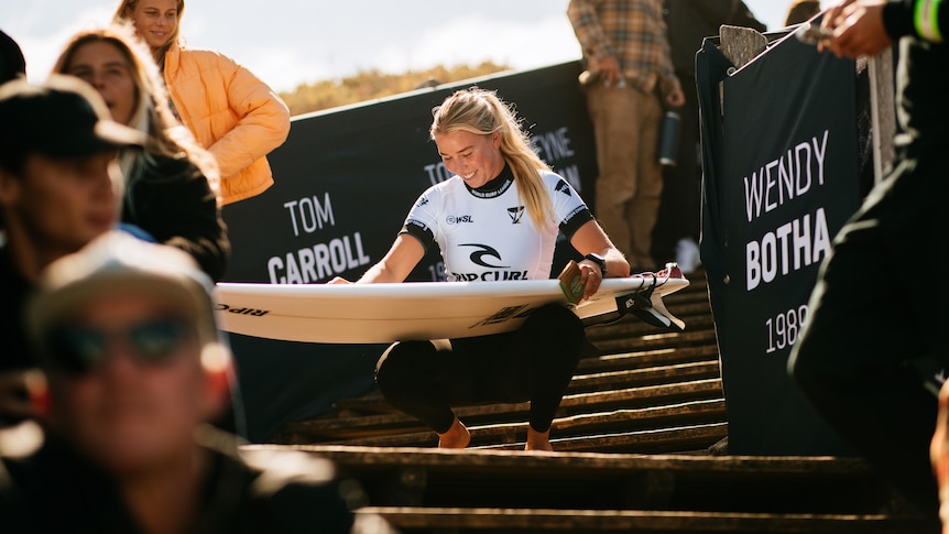 A young surfer smiles as she crouches on some steps holding her board before a competition.