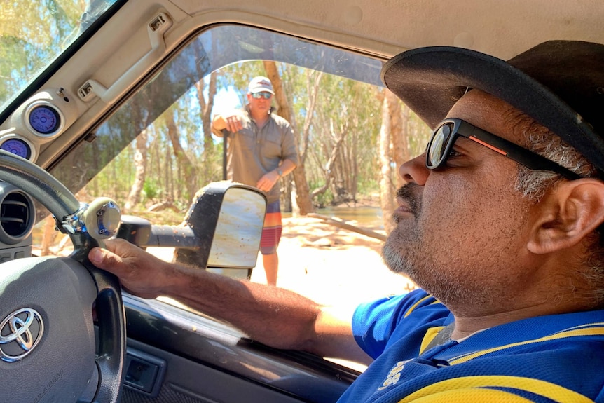 A man, wearing a hat and sunglasses, drives a four-wheel drive with a man looking on outside.