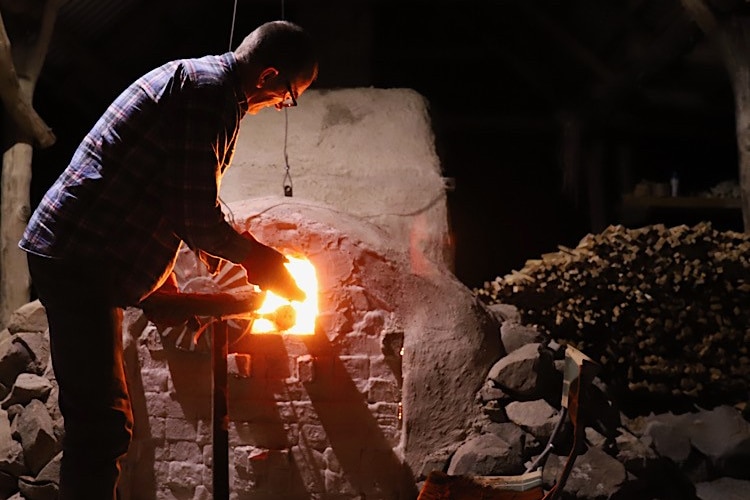 A man stands next to a woodfired kilm with a fire blazing, as he tends to pottery objects.