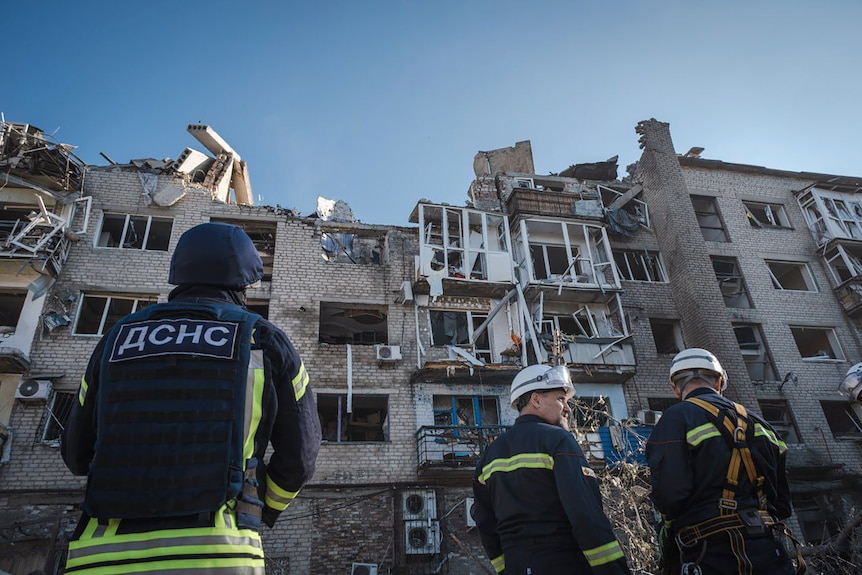 Trei bărbați purtând căști de înaltă vizibilitate stau în fața clădirii avariate 