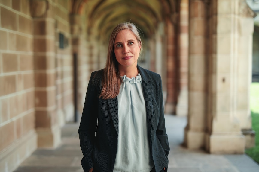 A woman stands in a covered sandstone walkway looking at the camera with a neutral expression.