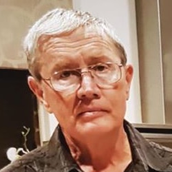 A tight head and shoulders profile shot of Ian Collett indoors wearing a brown shirt and spectacles.