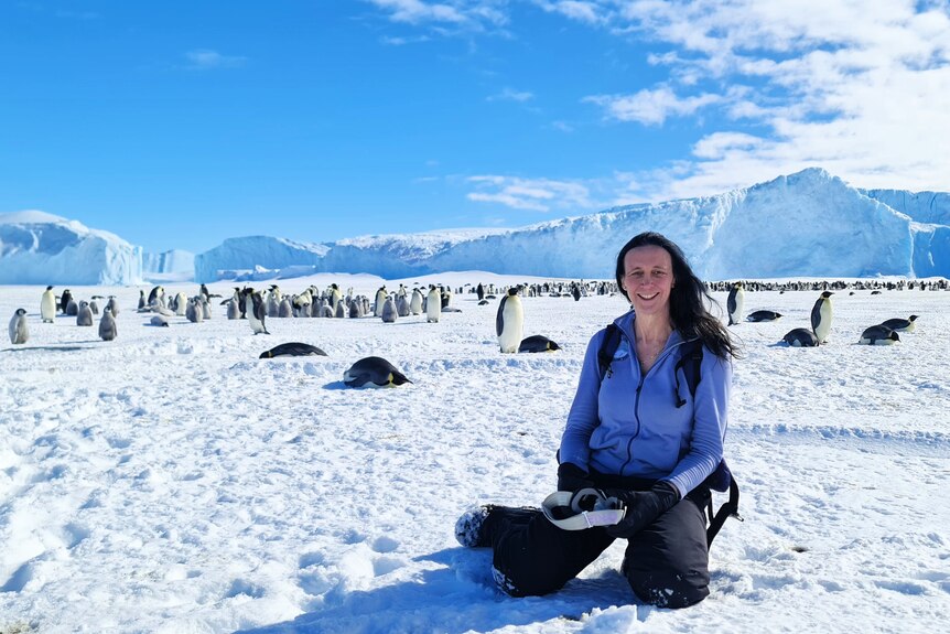 Cat Humphries squats down in the snow with penguins visible in the background