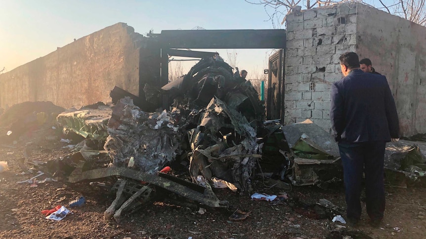 Two men are seen surveying the wreckage of a plane next to a brick wall. There are various objects around it.