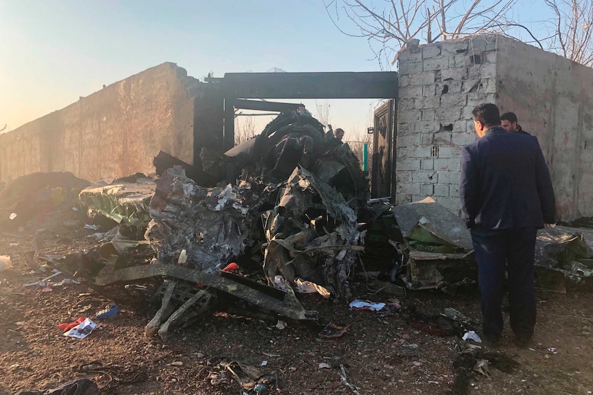 Two men are seen surveying the wreckage of a plane next to a brick wall. There are various objects around it.