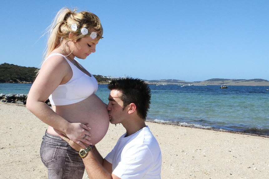 Young pregnant girl at beach scene with man kissing her round belly. 