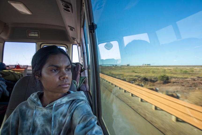 A young Indigenous woman sits on a mini bus, staring out the window to the outback landscape.