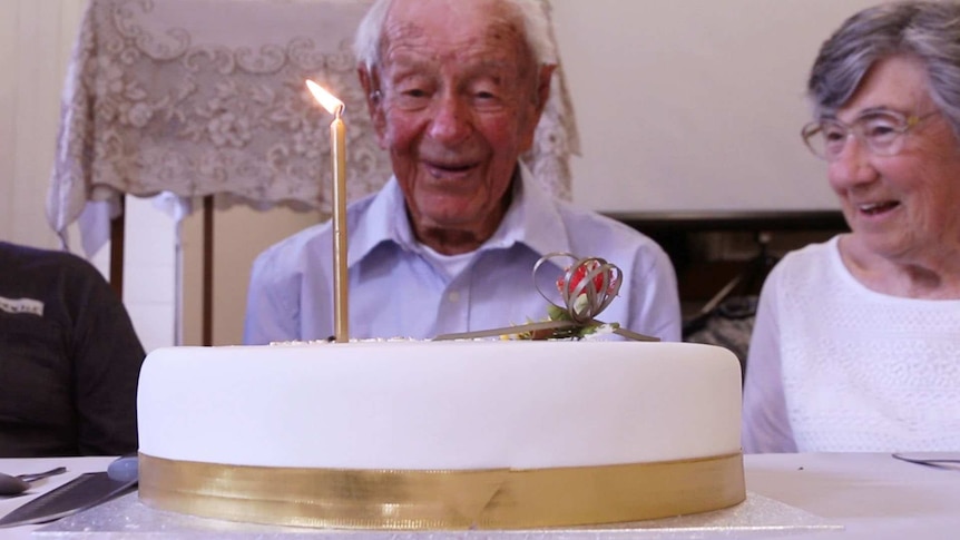 Birthday cake with candle in foreground, with elderly man sitting behind.