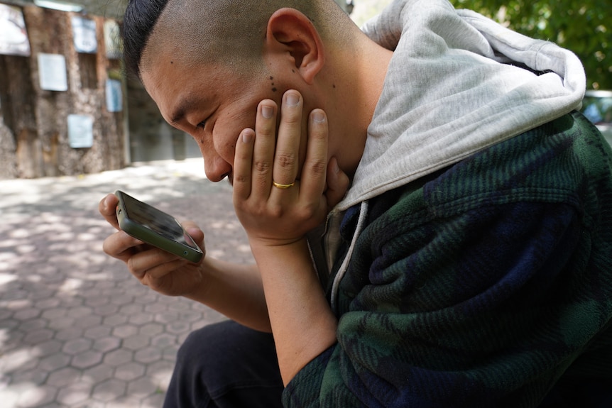 A young man looks at his phone while he holds his face in his hands and wears a jumper.