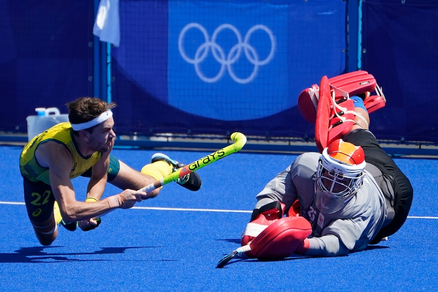 A man in green and gold dives with a hockey stick. A keeper is already on the ground.