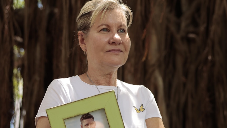 A woman looks away from the camera and holds a framed photo of a young man. 