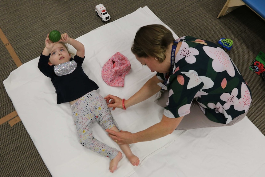 A little girl lays on the ground and holds a ball above her head while a woman measures her leg.