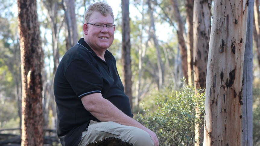 Peter Lacey sits on a log smiling and posing for a photo in bushland.