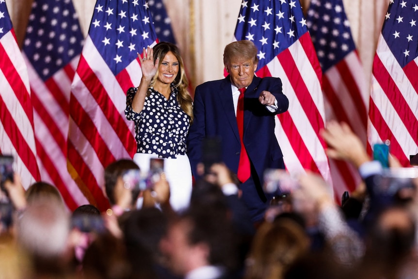 Melania Trump (left) and Donald Trump (right) dressed in a suit stand on stage smiling at a crowd of people.