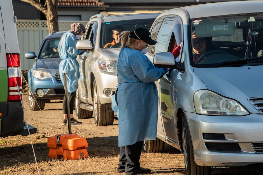 Two women stand at the window of two separate cars, with another car behind those two. They prepare to administer tests.