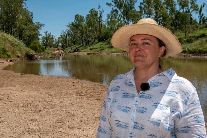 A woman in a hat smiles at the camera while standing on a riverbank