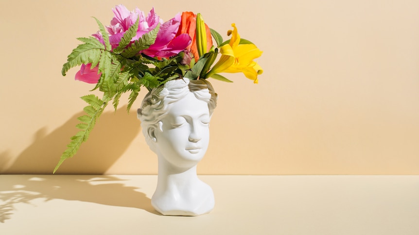 A bouquet of flowers flows from a vase that's the shape of a human head. the vase is white and its face is calm and smiling.