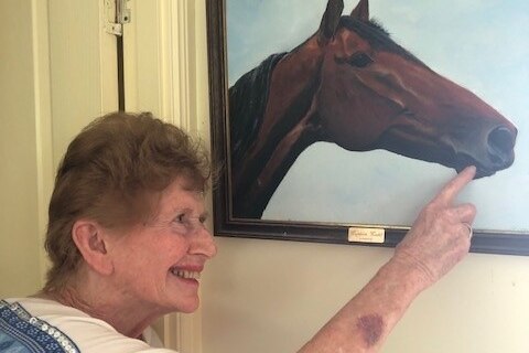 An older woman smiling and pointing at a painting of a brown horse. 
