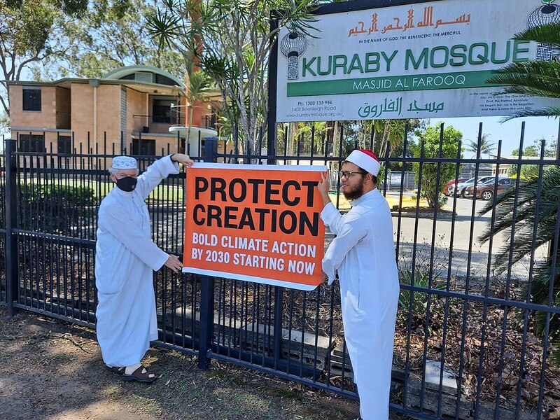 Two worshippers wearing white stand near a fence outside a mosque holding a sign that says 'protect creation'.
