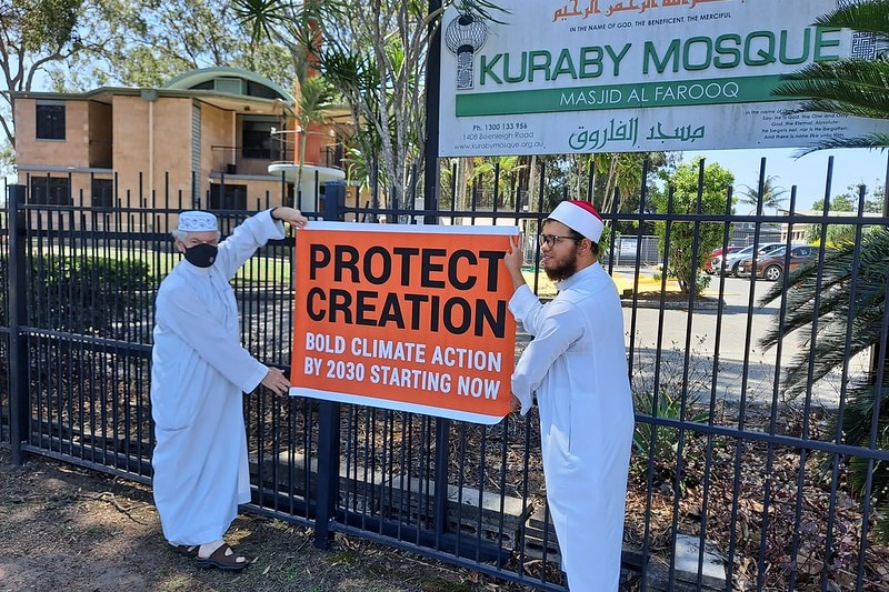 Zwei weiß gekleidete Gläubige stehen in der Nähe eines Zauns vor einer Moschee und halten ein Schild mit der Aufschrift „Beschütze die Schöpfung“.