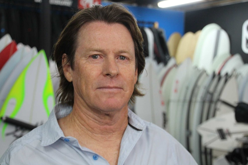 A head and shoulders shot of a middle aged man with longer hair and a collared shirt standing in a surf shop.
