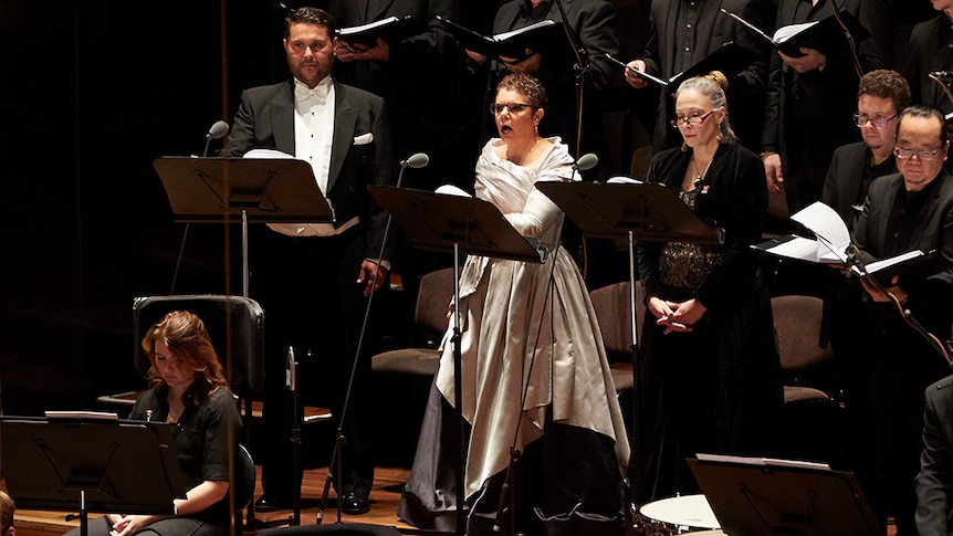 Composer Deborah Cheetham sings on stage amongst a choir.