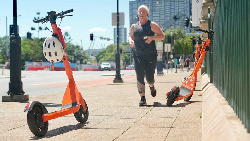 Woman jogging at Brisbane's South Bank navigates through orange e-scooters left parked on pedestrian walkways.