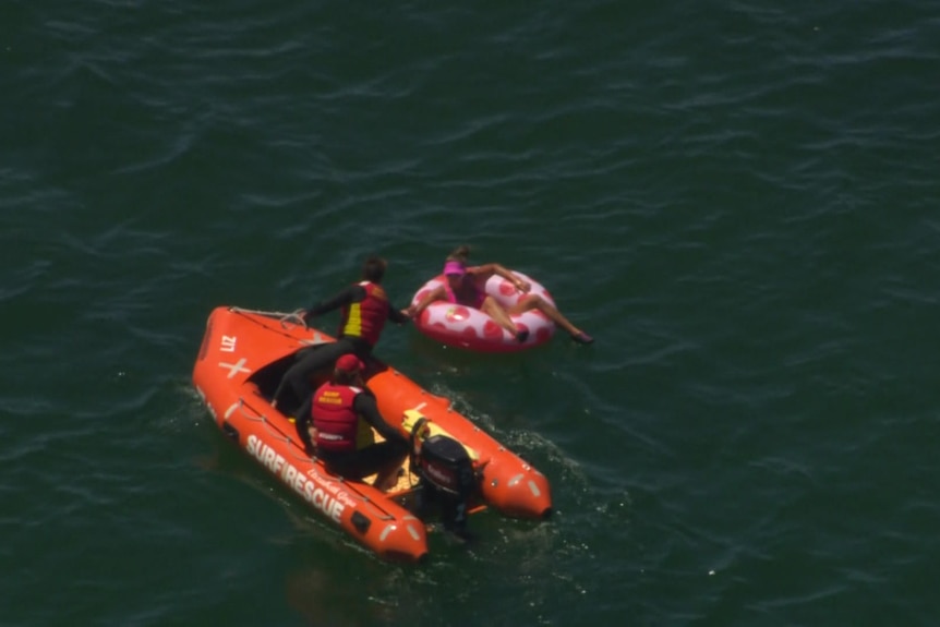 Two lifesavers in an inflatable boat reach towards a woman sitting on a pink polka dot inflatable ring.