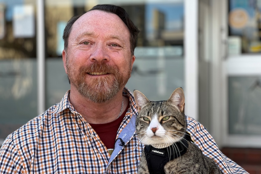Man wearing a checked shirt holding a cat that is wearing a harness. 
