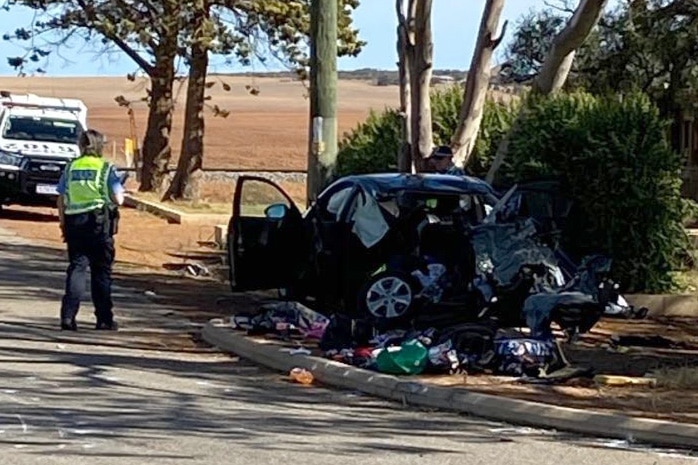 The wreckage of a sedan lies mangled on a dirt verge with debris strewn around it after a crash, with a police officer nearby.