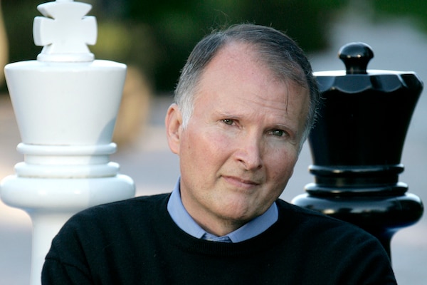 A man with grey hair and folded arms poses in front of large black and white chess pieces.