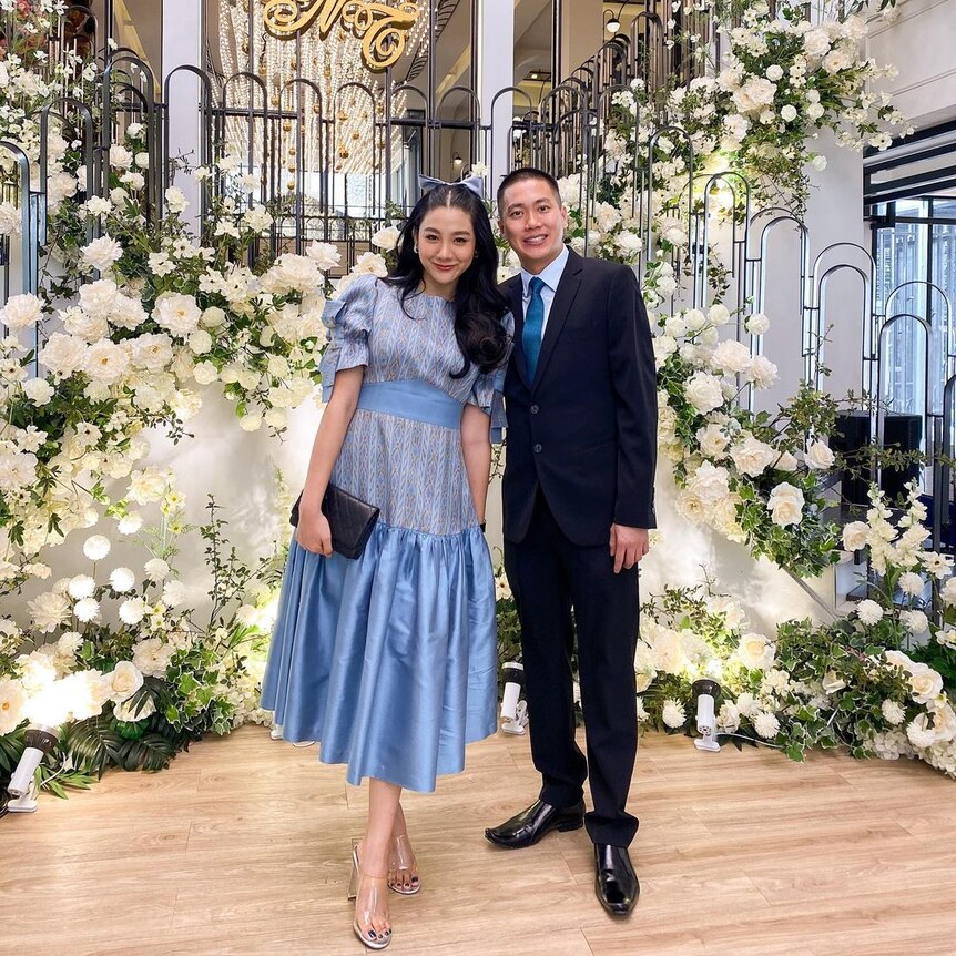 A Thai woman in a long blue dress and a man in a suit stand together surrounded by white flowers in a home