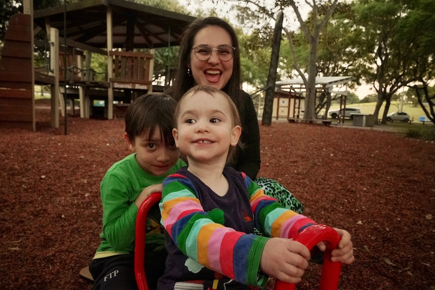 A mum and her two kids smile in a park.