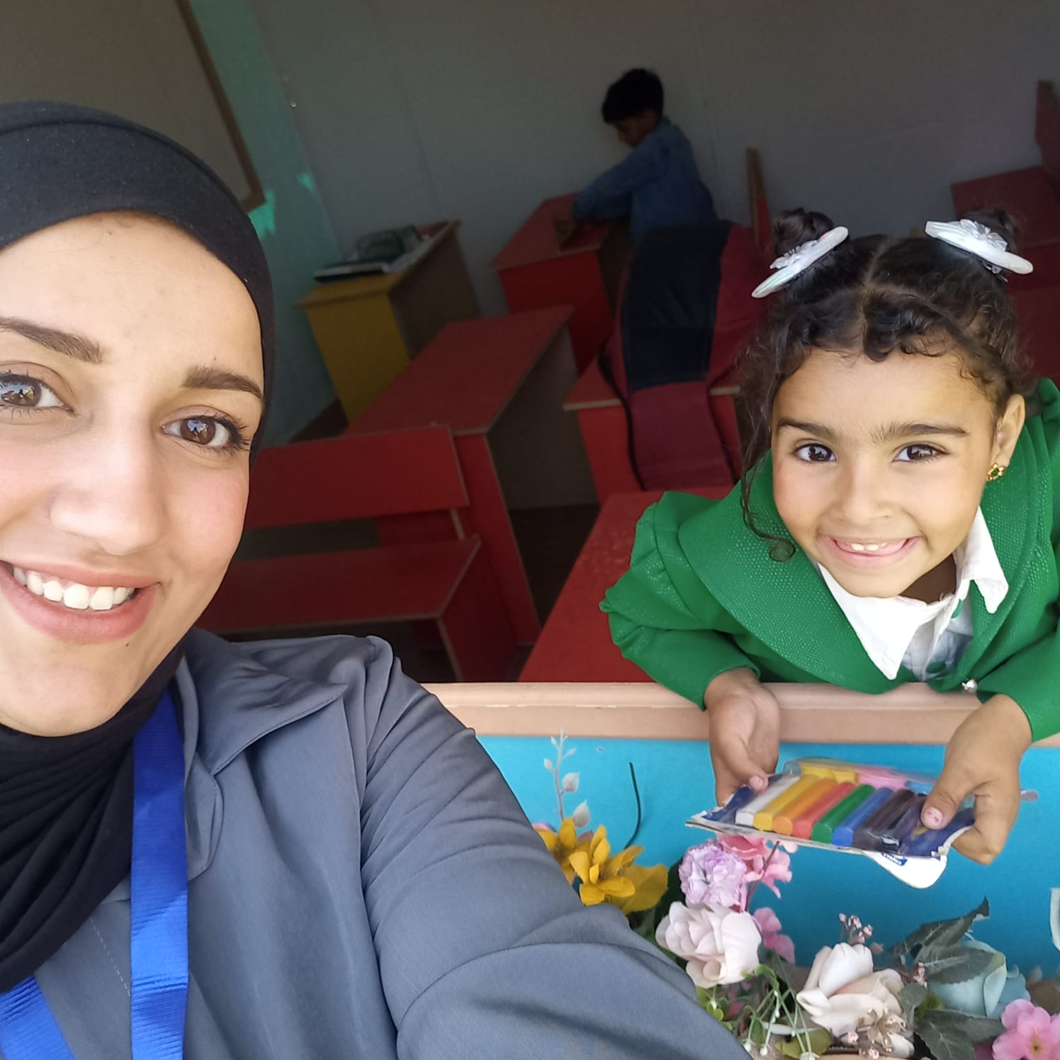 A Palestinian woman in a dark headdress and grey top smiling in a selfie next to a girl in a green top at a small table