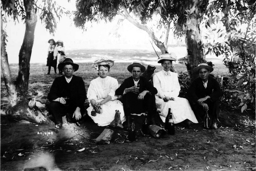 Black and white image of five young people sitting under gum trees in a dry creek bed, surrounded by bottles.
