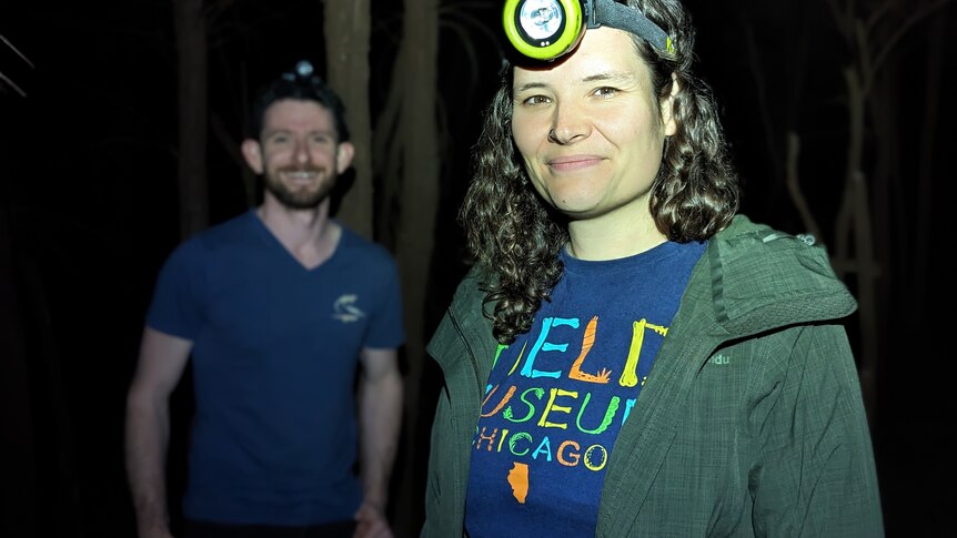 A man and woman wearing headtorches in the dark in a forest setting.