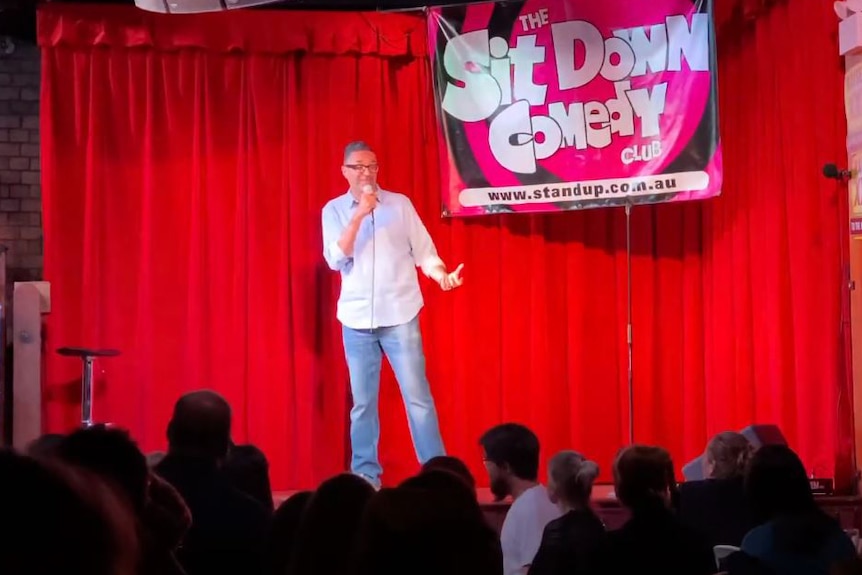 Vikesh holds a microphone as he stands on stage at a stand-up comedy event.