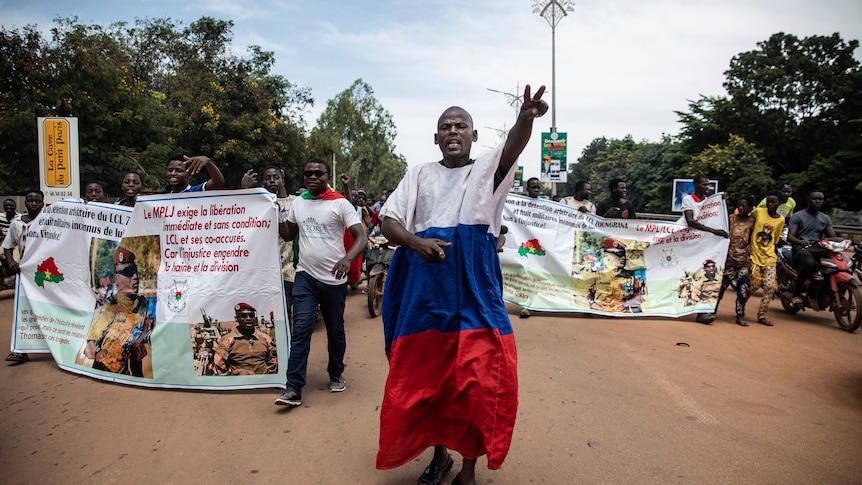 Protestors walk with banners with text. One man walks in front, making a peace sign with his hand wearing Russian flag colours