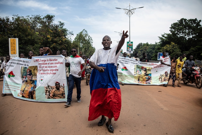 Protestors walk with banners with text. One man walks in front, making a peace sign with his hand wearing Russian flag colours