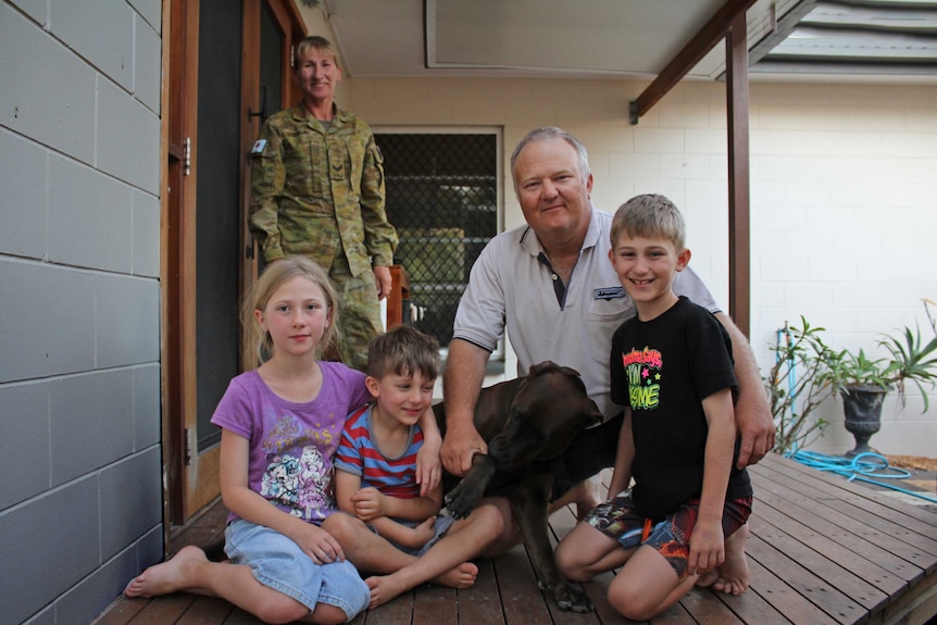 A man sits with his three children on their front porch, his wife is wearing Army uniform in the background