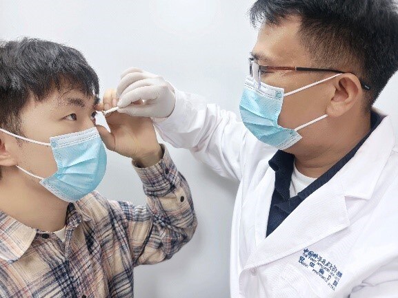 One person in gloves and a lab coat holding a small plastic contact lens applicator over the eye of another person.