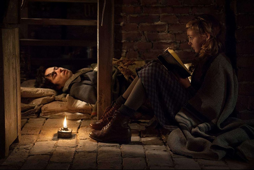 In an underground cellar a little girl reads a book by candlelight to a soldier lying down
