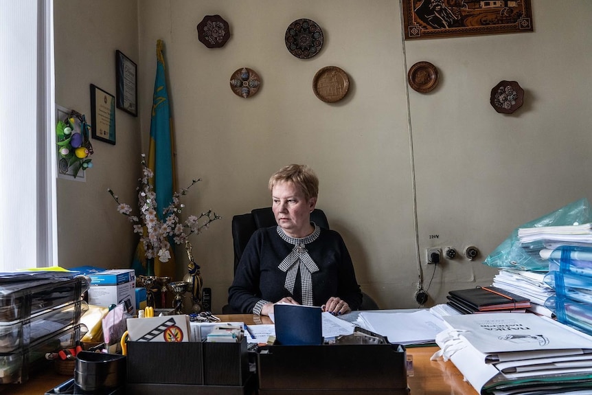 A woman with grey hair sits at her desk with flowers behind her.l